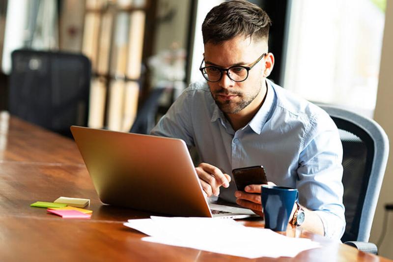 Serious man in glasses using laptop and phone with paperwork